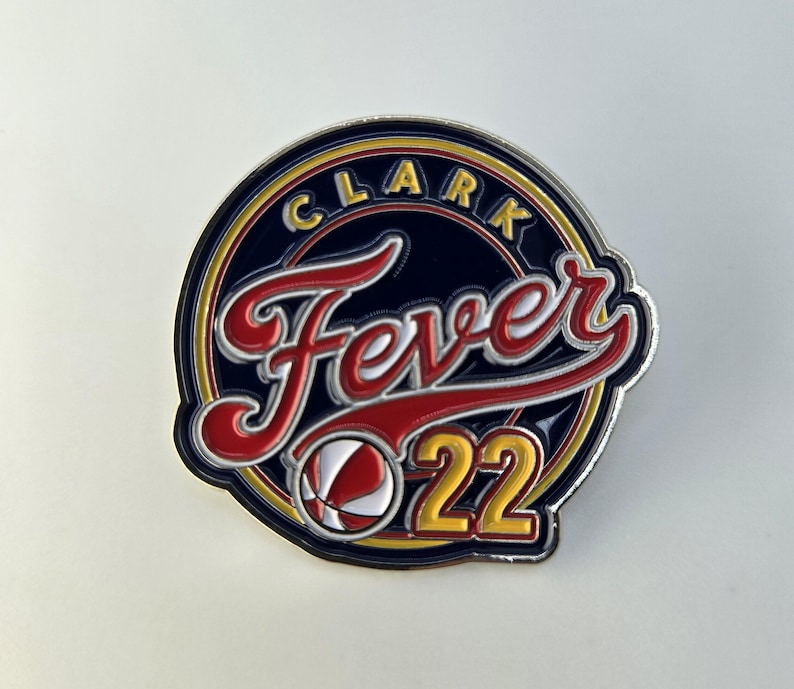 Puede incluir: Un pin circular azul y dorado con el texto "Clark Fever 22" en una fuente de letra blanca y roja. Un gr&aacute;fico de baloncesto est&aacute; en el centro del pin.