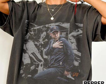 May include: A charcoal gray short-sleeve t-shirt featuring a large black and white graphic print of a person wearing a baseball cap and a hoodie, with their hand placed over their chest. The graphic includes a signature in orange ink.