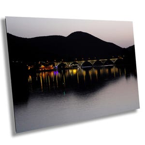 Op de afbeelding: Een nachtzicht van een brug verlicht met gele lichten, die reflecteren in het water. De brug bevindt zich voor een donker berggebied.