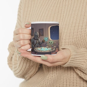 Op de afbeelding: Witte keramische mok met een Star Wars-thema illustratie. Het ontwerp toont de Mandalorian en Grogu die een bordspel spelen in een ruimteschip. De scène bevat een raam met uitzicht op een sterrennacht en gedetailleerde karakterafbeeldingen.
