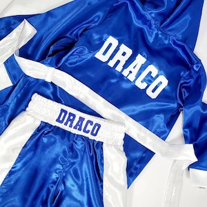 Puede incluir: Conjunto de bata y pantalones cortos de boxeo de sat&eacute;n azul con ribetes blancos. La bata tiene capucha y los pantalones cortos tienen cintura el&aacute;stica. Ambas piezas tienen el nombre "DRACO" impreso en letras blancas.