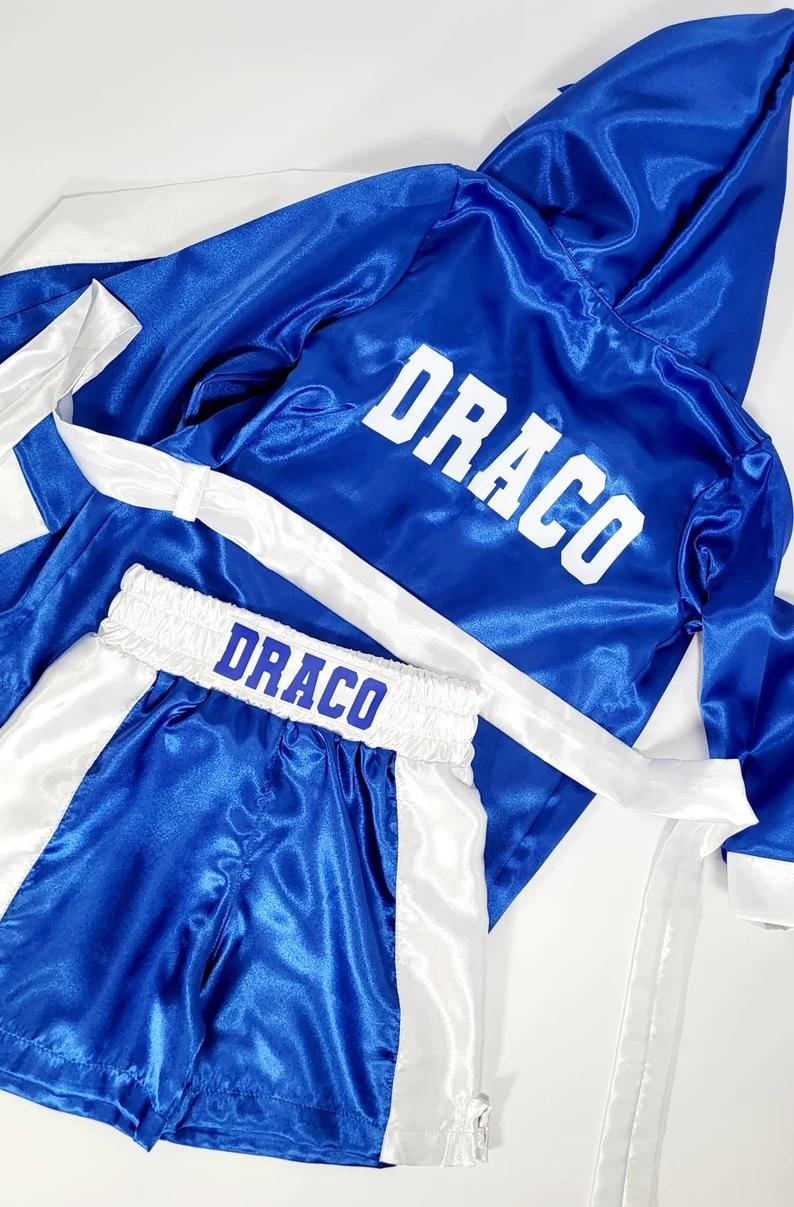 Puede incluir: Conjunto de bata y pantalones cortos de boxeo de sat&eacute;n azul con ribetes blancos. La bata tiene capucha y los pantalones cortos tienen cintura el&aacute;stica. Ambas piezas tienen el nombre "DRACO" impreso en letras blancas.
