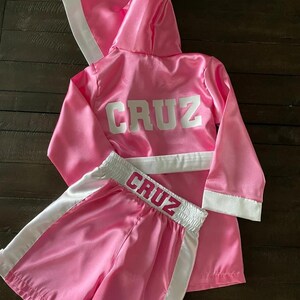 Puede incluir: Conjunto de bata y pantalones cortos de boxeo de sat&eacute;n rosa con ribetes blancos y el nombre "CRUZ" bordado en la parte posterior de la bata y en la cintura de los pantalones cortos.