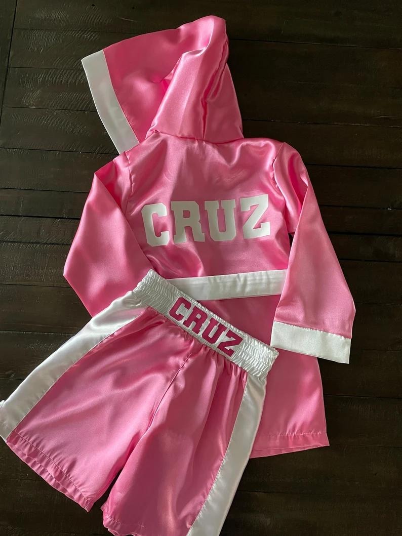 Puede incluir: Conjunto de bata y pantalones cortos de boxeo de sat&eacute;n rosa con ribetes blancos y el nombre "CRUZ" bordado en la parte posterior de la bata y en la cintura de los pantalones cortos.
