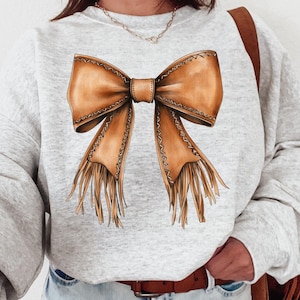 May include: A grey sweatshirt with a brown leather bow with fringe detail. The bow is centred on the front of the sweatshirt.