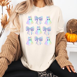 Puede incluir: Una camiseta blanca con un patr&oacute;n repetido de fantasmas iridiscentes que llevan lazos iridiscentes. Los fantasmas tienen ojos negros y un contorno azul claro.