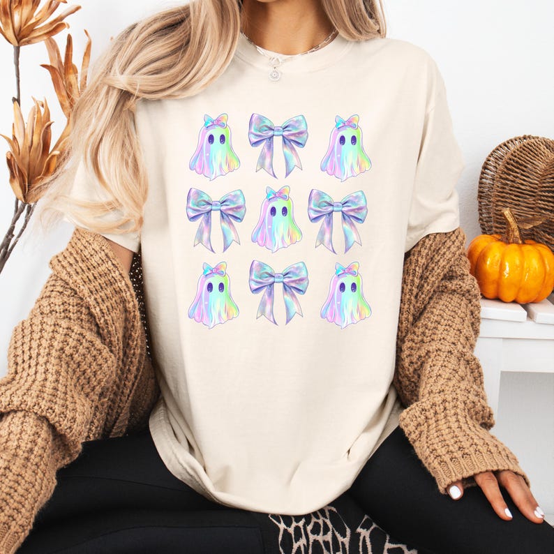 Puede incluir: Una camiseta blanca con un patr&oacute;n repetido de fantasmas iridiscentes que llevan lazos iridiscentes. Los fantasmas tienen ojos negros y un contorno azul claro.