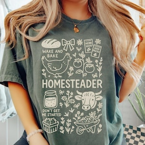 May include: A dark green t-shirt with a white graphic design featuring a loaf of bread, a chicken, tomatoes, and the word "HOMESTEADER". The text "WAKE AND BAKE" and "DON'T GET ME STARTED" are also included in the design.