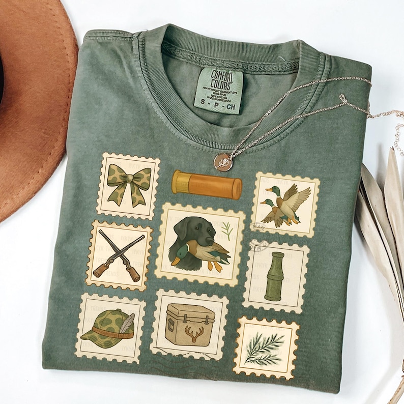 May include: Green Comfort Colors t-shirt with a hunting-themed stamp design. The stamps feature images of a bow, shotgun, dog with ducks, ducks in flight, a hunting hat, a cooler, a shotgun shell, a bottle, and a sprig of leaves. A silver necklace is draped across the shirt.
