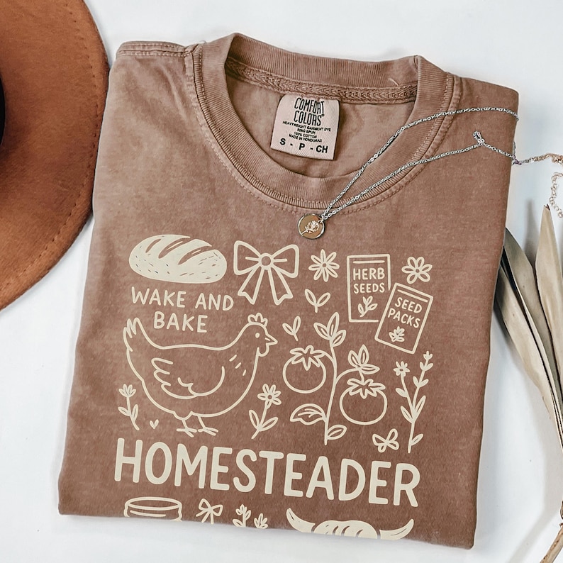 May include: A brown Comfort Colors t-shirt with a graphic design featuring a loaf of bread, a chicken, and the words "Wake and Bake" and "Homesteader." A silver necklace with a pendant is draped across the shirt.