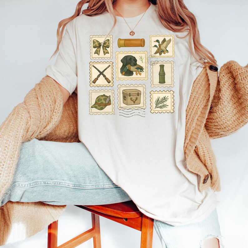 May include: Cream-colored t-shirt featuring a collection of postage stamp-style illustrations. The stamps depict hunting-related imagery, including a dog with a duck, crossed rifles, and a hunting hat. The shirt is worn by a person.