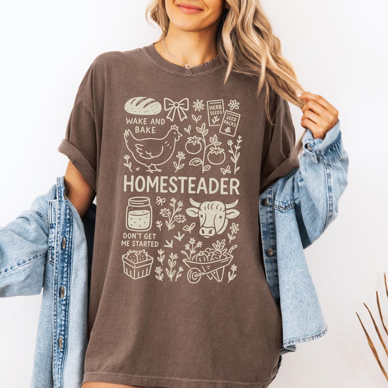 May include: Brown t-shirt with a cream-colored graphic featuring a loaf of bread, a chicken, tomatoes, a cow, and the word "HOMESTEADER". The text "WAKE AND BAKE" and "DON'T GET ME STARTED" are also included. The shirt has rolled-up sleeves.