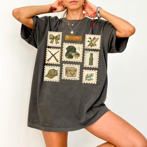 May include: Dark gray t-shirt featuring a collection of vintage-style postage stamps. The stamps depict hunting-related imagery, including a bow, ducks, a dog, a shotgun, a hat, and a cooler. The shirt has a relaxed fit and a slightly faded appearance.
