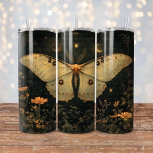 Puede incluir: Tres vasos de acero inoxidable con un diseño de una gran polilla verde claro con marcas marrones oscuras sobre un fondo oscuro con pequeñas flores amarillas. La polilla tiene una envergadura que cubre todo el vaso.