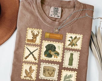 May include: Brown Comfort Colors t-shirt featuring a collection of hunting-themed postage stamp designs. The stamps depict a deer, ducks, a shotgun, a dog with a duck, a cooler, and other hunting-related imagery. A silver necklace is draped across the shirt.