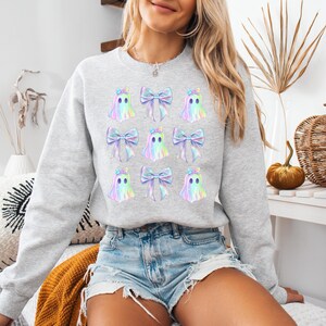 Puede incluir: Una sudadera gris con un patr&oacute;n de fantasmas iridiscentes y lazos. Los fantasmas tienen un solo ojo y est&aacute;n delineados en negro. Los lazos son en tonos de rosa, azul y verde.