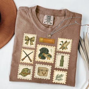 May include: Brown Comfort Colors t-shirt with a hunting-themed stamp design. The stamps feature images of a bow, shotgun shells, a dog with a duck, ducks in flight, a hunting hat, a cooler, and more. A silver necklace is draped across the shirt.