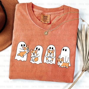 May include: Orange Comfort Colors t-shirt featuring a graphic of four white ghosts, each holding or accompanied by a corgi dog. The shirt has a crew neck and a small Comfort Colors tag. A gold necklace is draped across the shirt.