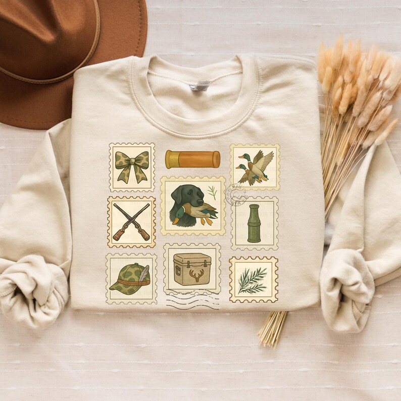 May include: Cream-colored sweatshirt featuring nine postage stamp-style illustrations of hunting-related items. The stamps include a bow, shotgun, dog with a duck, hunting hat, and other outdoor elements. The design has a vintage, outdoorsy theme.