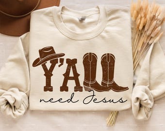 Könnte beinhalten: Cremefarbenes Sweatshirt mit dem Text "Y'ALL need Jesus" in Braun, mit Cowboyhut und Stiefel-Grafik. Das Design hat eine rustikale, ländliche Ästhetik. Das Sweatshirt ist ein lässiges, bequemes Kleidungsstück.