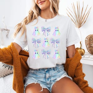 Puede incluir: Camiseta blanca con un patr&oacute;n repetido de fantasmas iridiscentes que llevan lazos. Los fantasmas est&aacute;n delineados en negro y tienen un degradado arco&iacute;ris.