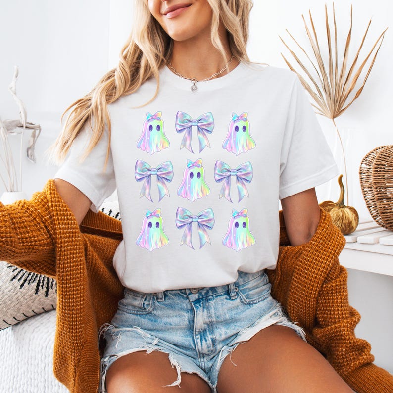 Puede incluir: Camiseta blanca con un patr&oacute;n repetido de fantasmas iridiscentes que llevan lazos. Los fantasmas est&aacute;n delineados en negro y tienen un degradado arco&iacute;ris.