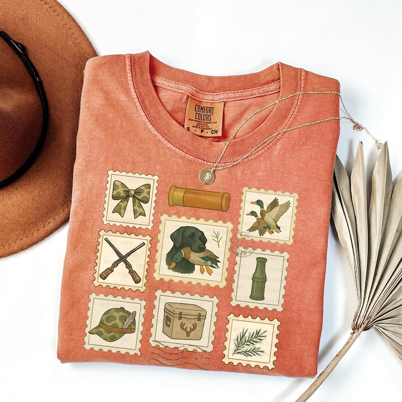 May include: A coral-colored Comfort Colors t-shirt featuring a collection of postage stamp-style illustrations. The stamps depict hunting-related imagery, including a bow, shotgun shells, ducks, a hunting dog, and a cooler. A gold necklace is draped across the shirt.