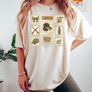 May include: Cream-colored t-shirt featuring a collection of postage stamp-style illustrations. The stamps depict hunting-themed images, including a bow, crossed rifles, a dog with a duck, and a hunting hat. The shirt has a relaxed fit.