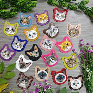 May include: A collection of embroidered cat face patches in various colors and patterns. Each patch features a detailed cat face with different fur colors and eye colors, surrounded by a contrasting border. The patches are arranged on a wooden surface.