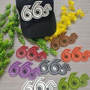 May include: A black baseball cap featuring a white and black "665" patch. Various colourful "665" patches in red, brown, orange, purple, black, white, and green are also shown. The patches have a stitched design.