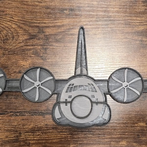 以下が含まれることがあります： 4つのプロペラと中央の胴体を持つ、グレーと黒の飛行機の模型。 木の背景に配置され、航空機のデザインと詳細を示しています。