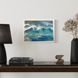 May include: A painting of a stormy sea with large waves crashing against each other. The painting is framed in white and is hanging on a white wall.