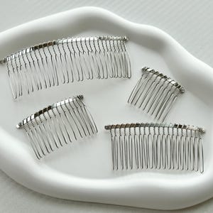 May include: Four silver hair combs on a white surface. The combs are different sizes and have a curved shape.