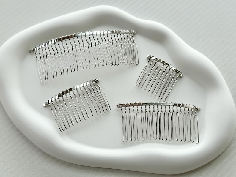 May include: Four silver hair combs on a white surface. The combs are different sizes and have a curved shape.