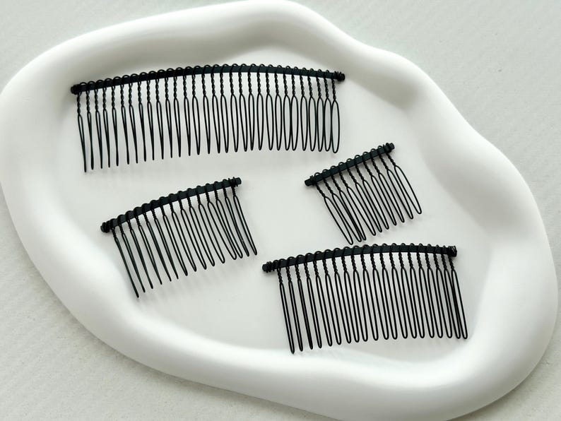 May include: Four black metal hair combs on a white surface. The combs are different sizes and have a curved shape.