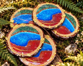May include: A set of four circular wood slice coasters featuring a resin inlay with vibrant blue, deep red, and green sections accented by gold leaf flakes. Each coaster displays a natural bark edge and a smooth, glossy finish on the top surface.