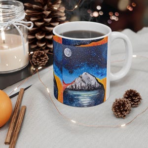 May include: White ceramic mug featuring a colorful landscape painting. The design includes a mountain, lake, and starry night sky with a full moon. The mug has a curved handle and is filled with a dark liquid.