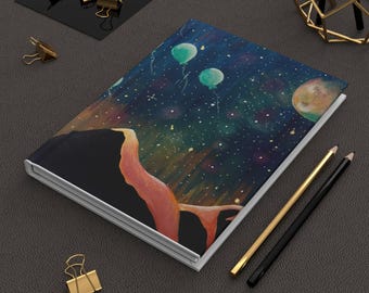 May include: A rectangular notebook featuring a dark blue night sky cover illustration with floating green balloons, a large orange planet, and scattered white stars. The notebook is positioned next to two pencils and gold binder clips.