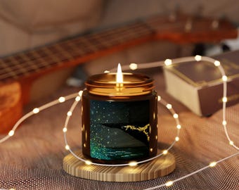 May include: A lit candle in a dark glass jar with a gold lid, set on a wooden coaster. The jar features a scenic design of a starry night over water. A string of fairy lights and a ukulele are in the background.