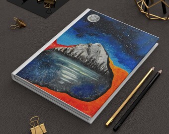 May include: A rectangular notebook featuring a cover illustration of a snow-capped mountain range and a forest reflected in a lake under a starry night sky with a full moon, set against a vibrant orange background, accompanied by two pencils.