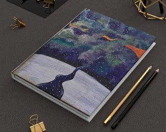May include: A rectangular notebook featuring a cover with a painted night sky scene, showing a dark blue starry expanse above a light gray landscape, accompanied by a gold pencil, a black pencil, and several gold metal binder clips.