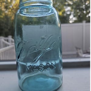 May include: A vintage light blue glass Ball Mason jar with a wide mouth opening. The front of the jar features the embossed cursive text Ball and the word MASON in capital letters. The glass has a translucent quality with a smooth cylindrical body.
