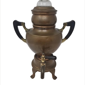Puede incluir: Percolador de café antiguo de cobre con tapa de cristal transparente y asas negras. La cafetera vintage tiene un grifo y se apoya sobre tres patas pequeñas. Un electrodoméstico de cocina clásico.