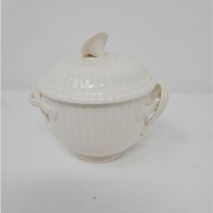 May include: Cream-colored ceramic sugar bowl with a lid and handles. The bowl has a ribbed design and a small, decorative handle on the lid. The handles are curved and appear to be woven. The sugar bowl is a classic kitchenware item.