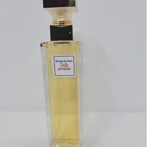 Puede incluir: Botella de perfume rectangular transparente con líquido dorado. La botella tiene un tapón dorado y negro y una etiqueta que dice "Elizabeth Arden 5th avenue". La botella está sobre una superficie blanca.