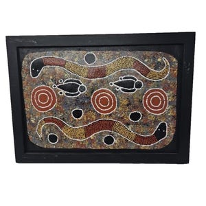 May include: Framed Aboriginal dot painting featuring two stylized snakes in shades of brown, yellow, and red, with black and white accents. The artwork includes circular and oval shapes, all set against a mottled background within a black frame.