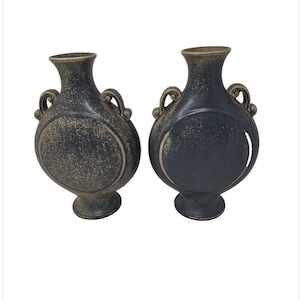 May include: Two dark blue ceramic vases with a speckled finish and a circular design. Each vase has a narrow neck, two handles, and a pedestal base. The vases are decorative and suitable for home decor.