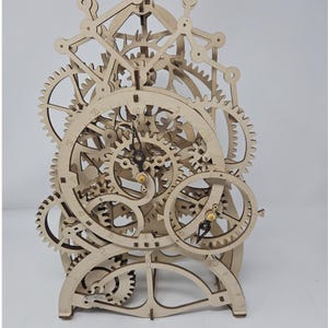 May include: A mechanical clock model constructed from light-colored wood, featuring an intricate assembly of exposed gears, cogs, and levers. The clock face displays numerical markings, and black hands are positioned at the center of the mechanism.