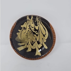 Puede incluir: Pequeño plato redondo de cerámica con interior negro y diseño dorado y negro arremolinado. El borde es marrón texturizado. El plato, probablemente hecho a mano, es ideal para servir pequeñas porciones de comida o como objeto decorativo.