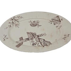 May include: An oval-shaped ceramic platter with an off-white base color and brown transferware patterns. The design features scattered bamboo sprigs, a small fan-like motif, a diamond shape, and a central arrangement of folded ribbons and floral patterns.